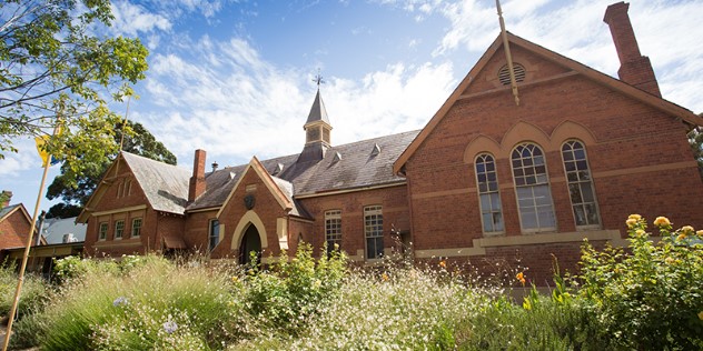  A brick, Victorian style home with two wings and a small turret, framed by grass and native flowers.