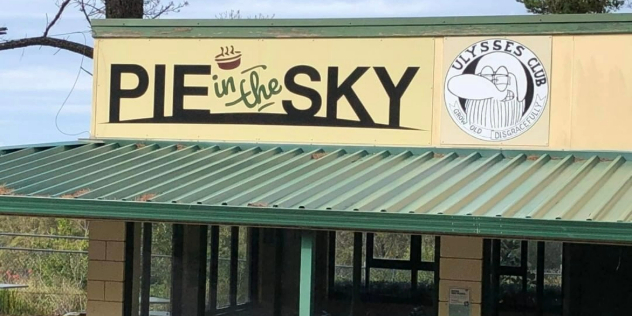 Flat topped cafe with tables outside with a sign at the top saying Pie in the Sky and a cartoon of a man with a beard. 