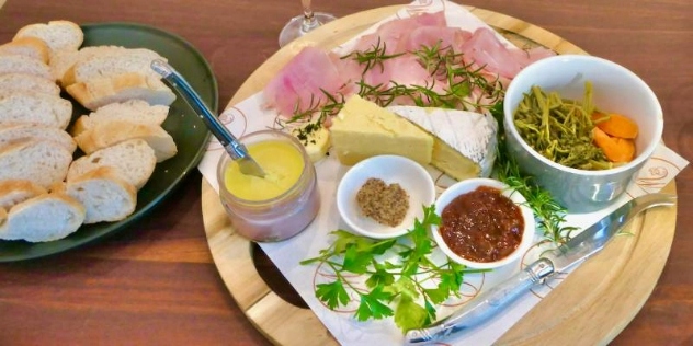  A wood platter of meat, cheeses and pickled vegetables, next to a plate of bread slices on a wood table.