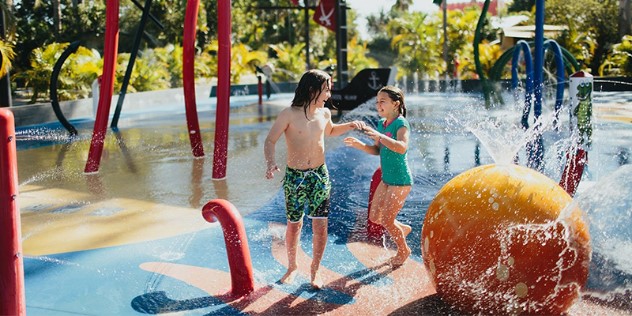 A boy and girl play among colourful water features by a pool on a sunny day. 