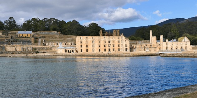 Port Arthur historical military buildings made of beige stone, as seen from a boat as it enters the port under a blue sky.