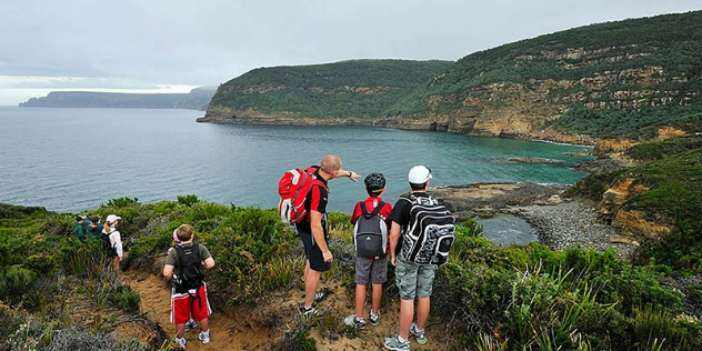 A group in backpacks walk a grassy cliff path down towards a bay, with green heads of land extending along the shore.