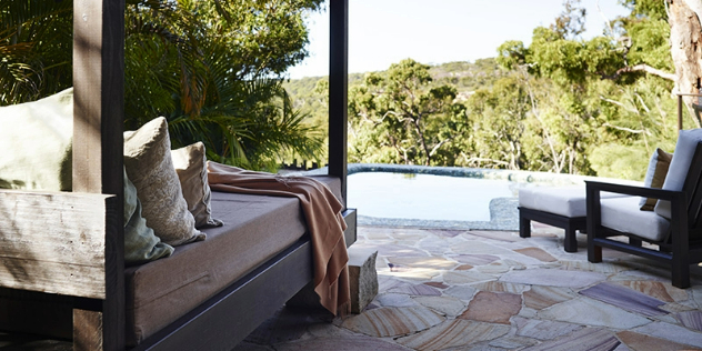  A four poster day bed with pillows and chairs next to a spa bath on the corner of a veranda, in a sunlit treetop. 