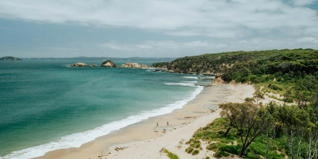 A curved nearly white sand beach ending with lush green shores seen from a distance, with two small hikers visible.