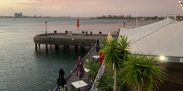  A rounded pier jutting out from a boardwalk along a lake, lined with umbrellas and restaurants.