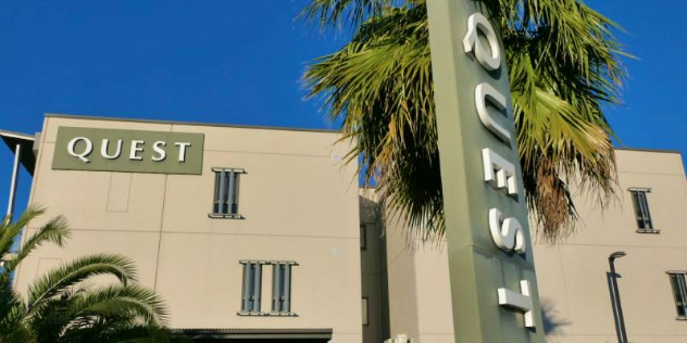  The facade of a rectangular building with Quest written along the side, framed by palm trees.