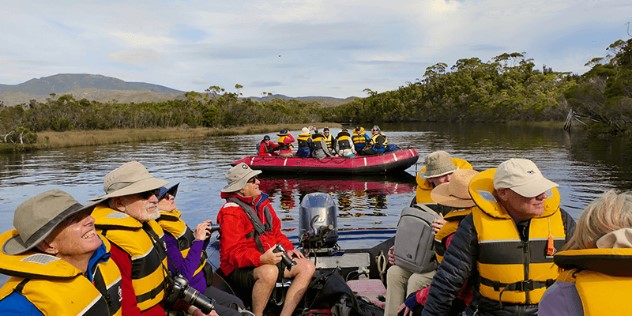 Two red and black inflatable rafts filled with tourists in yellow lifejackets drifts through a brown-water inlet, surrounded by mangrove trees on a sunny day. 