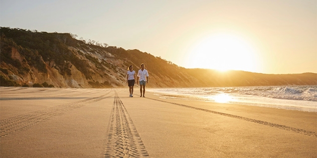 a man and a woman wearing shorts and white shirts walk along the beach following four-wheel-drive tracks at sunset