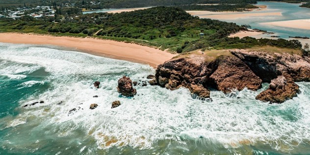 A long sandy beach, and treed peninsula leads to a rocky outcrop, surrounded by smaller rocks and sea foam, on a sunny day.