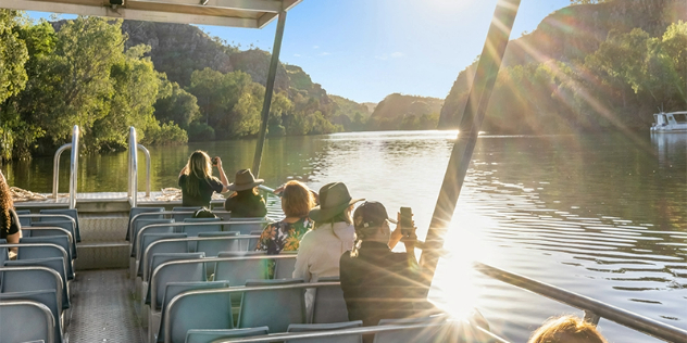 Sun flashes off the water as a tourboat goes along a river surrounded by tall green hills and mangroves. 