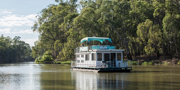 A rectangular houseboat with a veranda on the roof, gliding down a tree lined river.