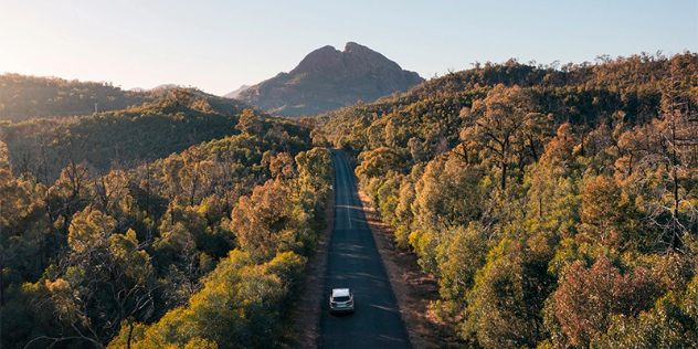 A car travelling a paved, forest lined road lit by sunlight, with a mountain peak rising over the horizon.