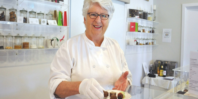 A woman with short white hair and glasses in a chef's jacket, holding a plate of chocolates at cafe counter.