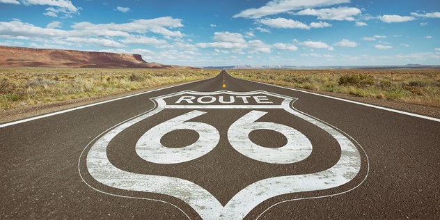 Close up of a road with Route 66 painted in white on the pavement, cutting through a panoramic view of a scrubby desert and rock formation.