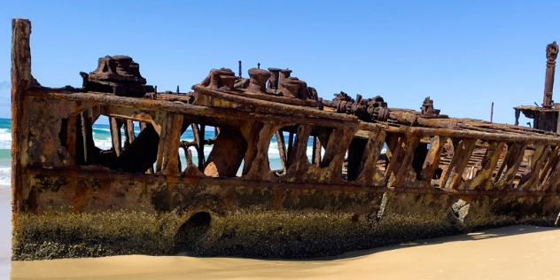  A rusted ship bottom, sitting on sand under blue sky.