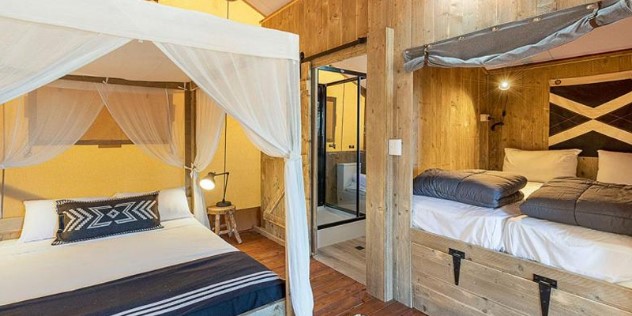 A room with a four poster canopy bed, hung with white fabric and two single beds mounted next to it, inside a long rectangular cubby hole built into a natural wood wall. 