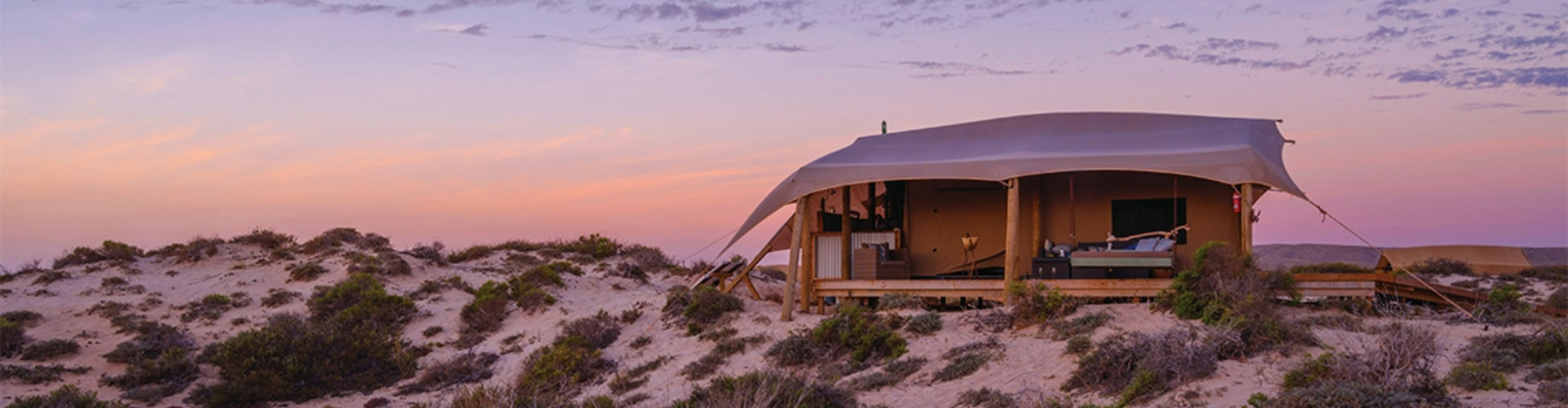 sal-salis-glamping-tent-exmouth-wa-1920x500 A wood-frame tent with a tarp on top and one side open revealing a queen bed, all sitting on a sandy dune, lit by a sunset.