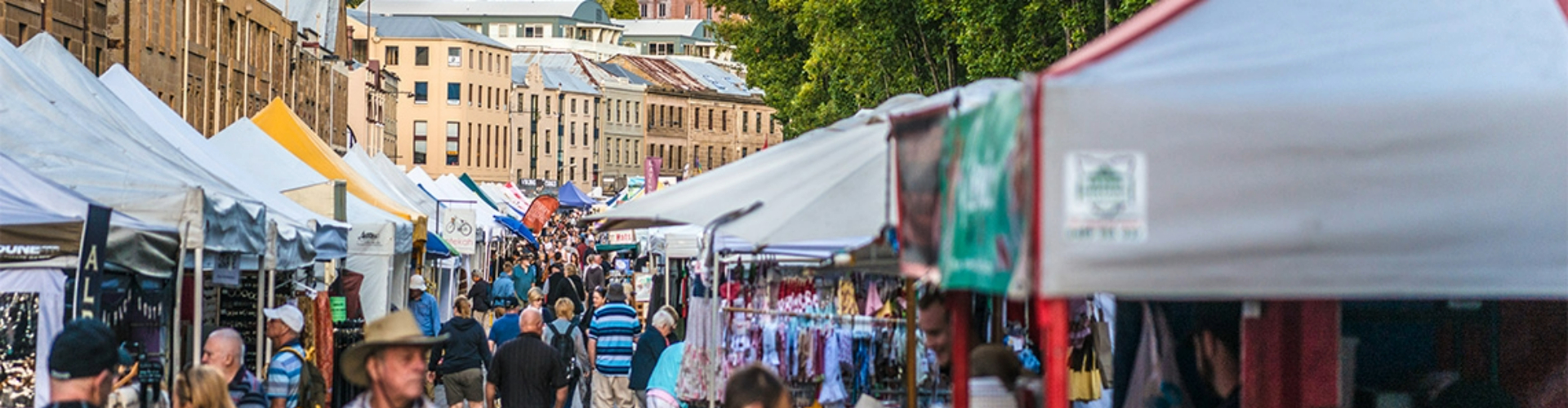 salamanca-markets-hobart-tas-1920x500 Crowds walk through market tents, with a row of historic buildings up the street behind them.