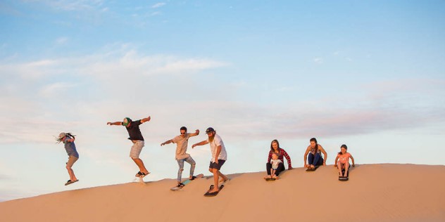 A mother with teens and small kids, all sitting or standing on long boards preparing to slide down a sand dune.