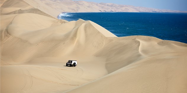 A single SUV appears small driving across huge sand dunes that back up onto deep blue sea.