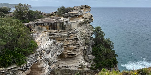 A rocky outcrop of sandstone-cliffs against a dark ocean horizon and grey sky, the cliff has a largely flat top, with similar flat layers stacked underneath, surrounded by large trees clinging along the cliff's edges.