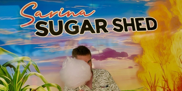 man holding pink candy floss on a stick stands in front of mural that says Sarina Sugar shed