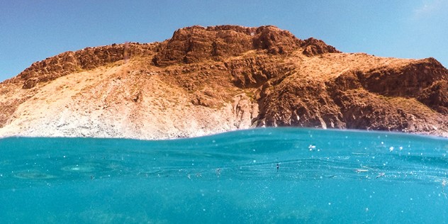 A red rocky reef juts up from bright blue water as seen from the perspective of a swimmer, framed by blue sky.