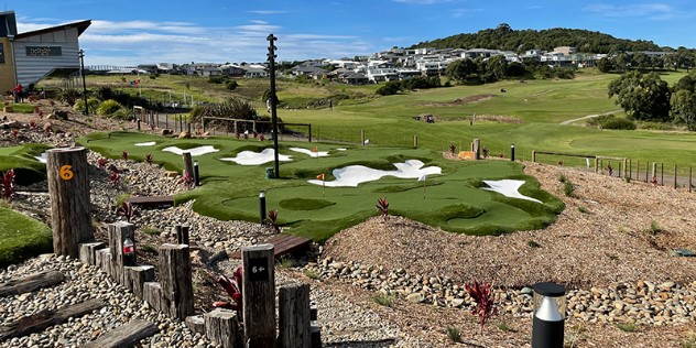 Part of a mini golf course, with greens and white sand traps among small red flags, with a town visible on green hills in the background.