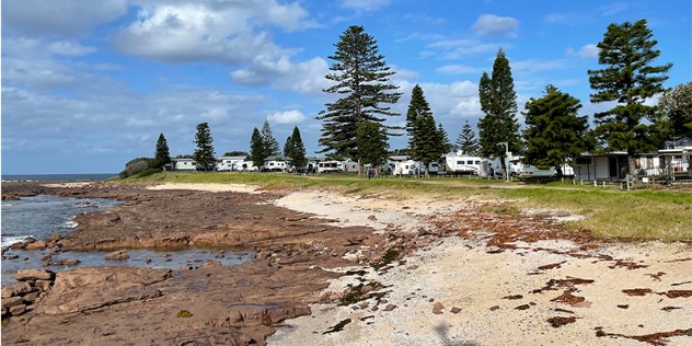 A campground along a rocky beach with trailers parked on the grass among trees in the background, under blue sky.