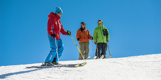  Three skiers in hats at the top of a white snowy hill framed by blue sky.