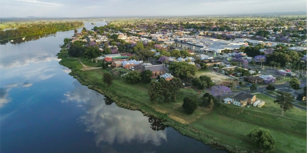 A small, low-lying town spread out along a river bend. There is no sand or rocks on the river shore, instead from the water to around the town and into the distance is all lush, green grass with patches of trees.