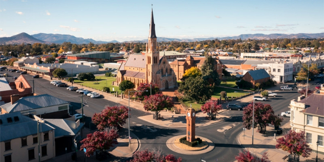 A small town seen looking down over the town centre, at low-lying, older style commercial buildings, a small roundabout with a clock tower and a steepled church, framed by trees and rolling hills in the distance. 