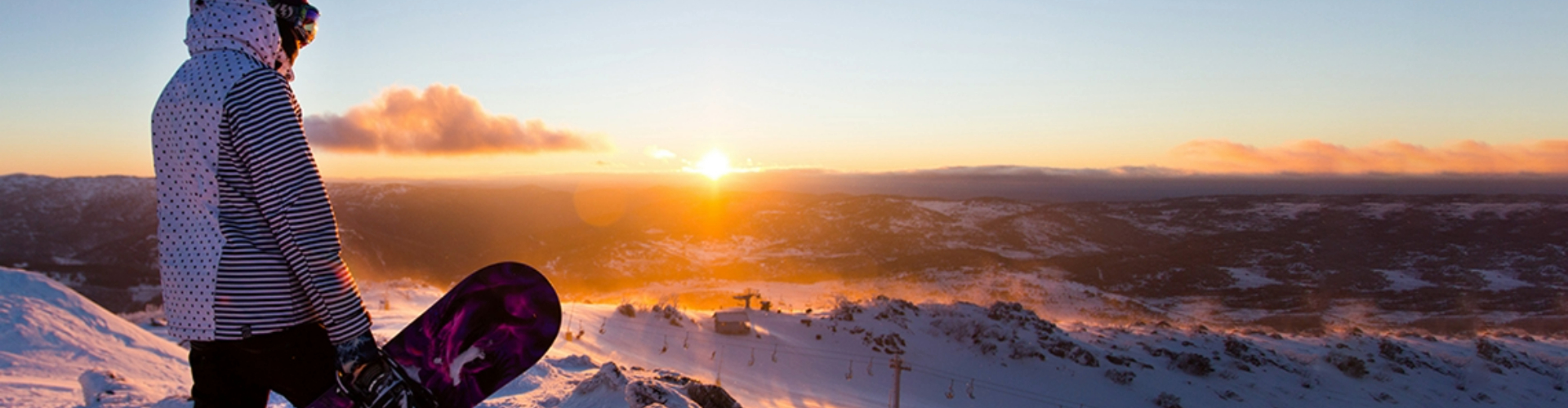 snowboarding-snowy-mountains-nsw-1920x500 A snowboarder in a hoodie on a mountaintop, looking over peaks bathed in orange sunset light.