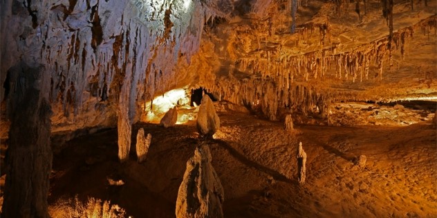 The interior of a cave lit yellow from a single light source, illuminating a cave ceiling full of stalagmites hanging down, while the cave floor is sandy and relatively smooth.