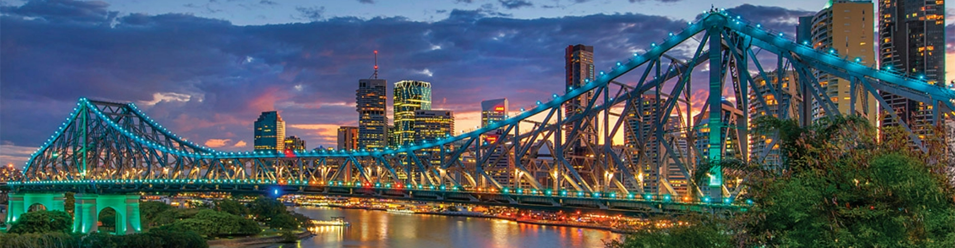 story-bridge-brisbane-skyline-1920x500 A double-steepled bridge, with girders lit with blue lights over a river reflecting glowing city skyscraper lights.