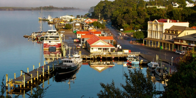 A small town harbour at golden hour, boats moored along the board walk, with a street and colonial style buildings visible on shore.