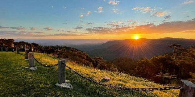 A cliffside lookout view of a sunset over the Blue Mountains on the horizon. past a deep forested valley.