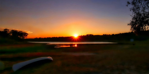  A treed lakeshore, with a canoe laying in the grass, lit orange by a sunset. 