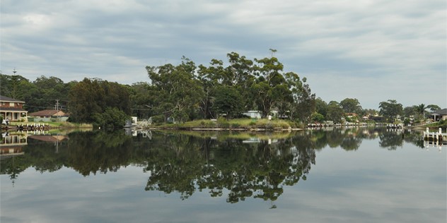 Still water of a canal on an overcast day, the water reflection mirroring the trees and few houses on the opposite, grassy shore. 