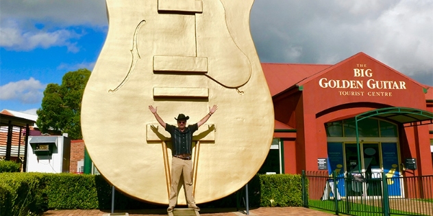 man in black stetson hat with his arms outstretched dwarfed by huge golden guitar behind him that also towers over the tourist centre behind it