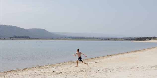 A teen boy in black shorts runs down a near-white sand beach towards the water, with green hills in the distance on an overcast day.