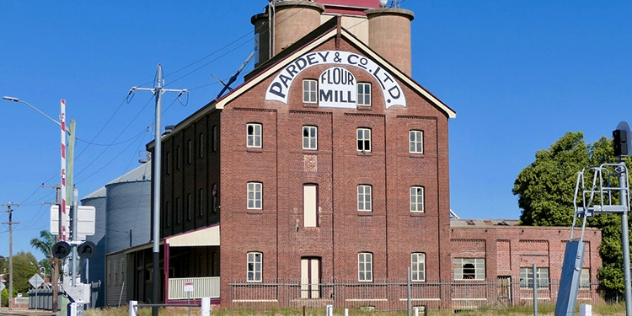  A brick mill along a train track framed by blue sky, Pardey and Co. Lt. Flour Mill is written in vintage script above the mill windows.