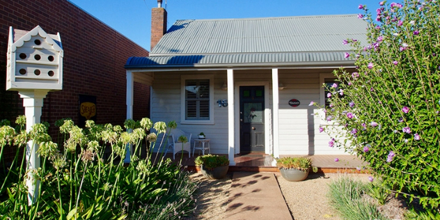  A small white cottage house with a birdhouse and flower bushes along its front walkway.