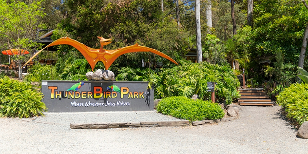 A concrete path leads to a sign saying ThunderBird Park with a large orange pterodactyl outstretched over it and a stairs leading to a forested area.