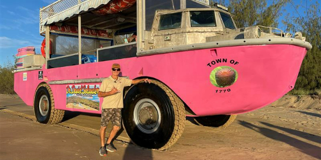  A man leans against the wheel of a bright pink amphibious boat with a metal canopy, on a sandy, sunlit shore.