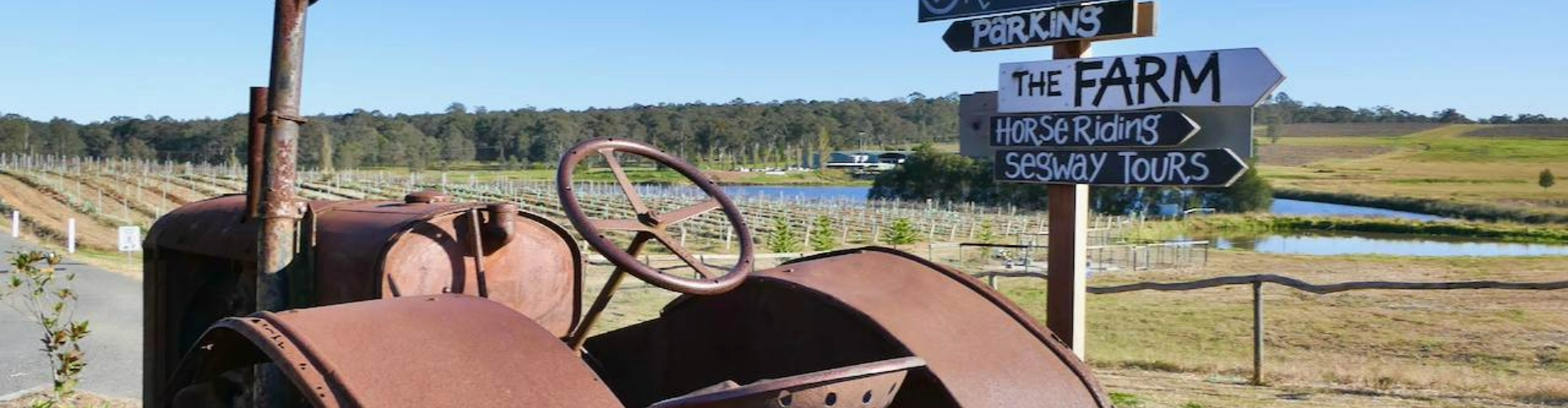 tractor-at-hunter-valley-resort-nsw-1920x500 An old, rusted tractor next to a sunny vineyard and a signpost with signs for things like horseriding and segway tours.