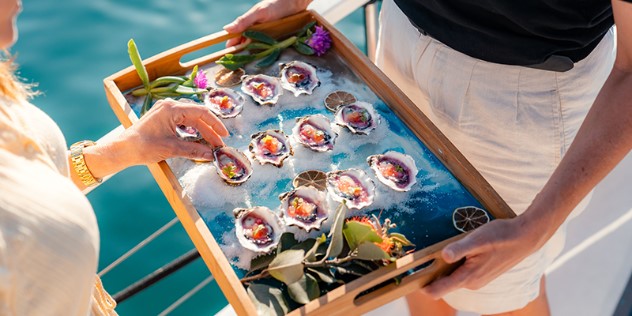 A wooden tray of twelve garnished oysters, being offered to a blond woman on a boat. 