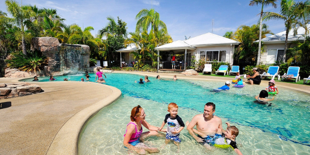  Families play and relax in a landscaped resort pool area, surrounded by palm trees and cabins.