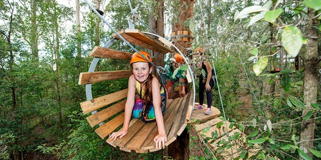 happy girl in orange hard hat and rainbow-coloured leggings pauses inside cylinder suspended from the treetop climbing course