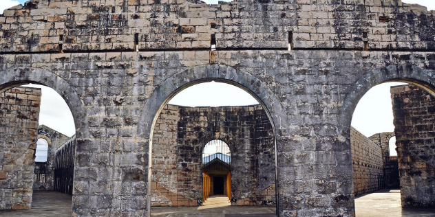 A historic, stone prison wall without a roof, with three curved passthroughs, behind which is a door-less entry and more stone walls, under blue sky.